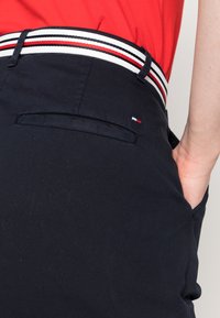 Pantaloni navy con una cintura a contrasto a righe nei colori rosso, bianco e nero. Include una tasca posteriore e un dettaglio del logo del brand ben visibile.
