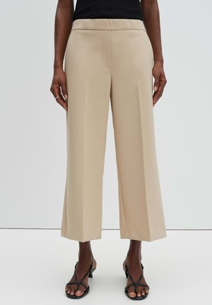 Frau trägt beige weite, verkürzte Hosen und schwarze, geschnürte Absatzsandalen, steht vor einem schlichten weißen Hintergrund.