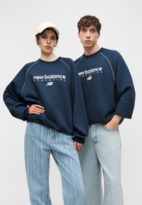 Zwei Models tragen dunkelblaue New Balance Athletics Sweatshirts und hellblaue Jeans und stehen nebeneinander vor einem schlichten Hintergrund.
