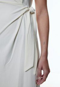 Weißes Kleid mit einem Knoten-Detail an der Taille, aus weichem Stoff gefertigt. Nahaufnahme einer Hand, die auf dem Kleid ruht, mit einem Tattoo am Handgelenk.