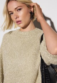 Hellbeiger, flauschiger Pullover mit strukturierter Optik, Rundhalsausschnitt und sanftem Schimmer. Das Modell trägt auffällige schwarze Ohrringe und hält eine schwarze Handtasche.