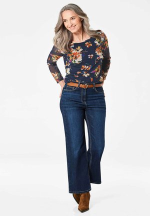 Een bloemen top met lange mouwen op een model, gecombineerd met wijde, donkere spijkerbroek en een leren bruine riem. De outfit heeft levendige bloemmotieven.