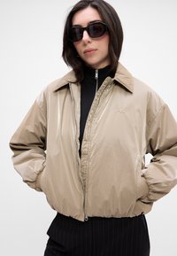 Beige padded jacket with a glossy finish, zip closure, and elastic cuffs. Features a classic collar and CK logo embroidered on the chest.