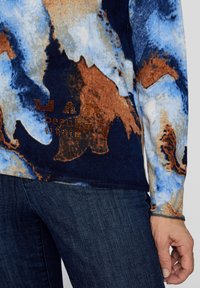 Langärmliges Oberteil mit einem blauen, braunen und beigen abstrakten Muster, einer strukturierten Oberfläche und 'beauty' mit Nieten bedrucktem Schriftzug auf der Vorderseite.