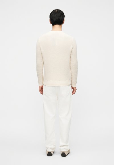 Ανοιχτό μπεζ πλεκτό πουλόβερ με υφασμάτινη υφή, εφαρμοστά μανίκια και τελείωμα. Συνδυάζεται με λευκό παντελόνι και μπεζ παπούτσια.