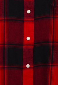 Chemise à carreaux rouge et noire avec fermeture à boutons, boutons en plastique blanc et texture de tissu doux avec des lignes horizontales et verticales visibles.