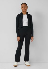 Schwarze, kurz geschnittene Jacke mit Reißverschluss über einem weißen Oberteil, kombiniert mit schwarzen Schlaghosen und weißen Turnschuhen mit beiger Sohle. Einfaches und figurbetontes Design.