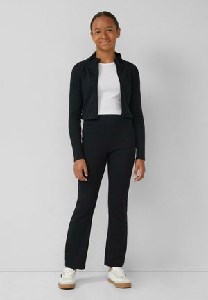 Veste zippée noire cropped sur un haut blanc, associée à un pantalon noir évasé et des baskets blanches avec une semelle beige. Design simple et ajusté.