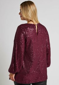 Burgundy paljettopp med långa puffärmar, rund halsringning och en nyckelhålöppning bak, med en slät, texturerad yta.