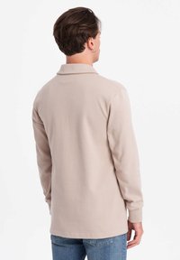 Beige långärmad polo med krage, tillverkad av mjukt tyg. Har en rak fåll och ribbade manschetter. Bakvyn visar en enkel design.