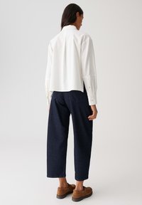 Vrouw met lang donker haar, gekleed in een losse witte shirt, donkere wijde jeans en bruine suède schoenen, staand tegen een effen achtergrond.