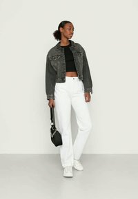 Mulher em pé com calças brancas, top preto cropped, casaco de denim preto desbotado, ténis brancos, segurando uma pequena bolsa preta com detalhe em corrente.