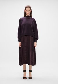 Ljubičasti pleteni pulover s okruglim izrezom, uparen s lepršavom tamno ljubičastom midi suknjom. Crne sandale s remenom oko gležnja upotpunjuju izgled.