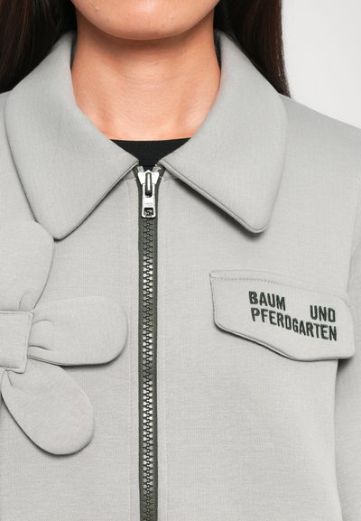 Grå zip-up jakke med en stor sløjfedetalje på venstre side, ribbet tekstur, en grøn lynlås og et mærke med "BAUM UND PFERDGARTEN."