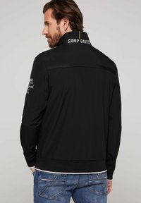 Sort zip-jakke med høj krave, der har "CAMP DAVID" broderet på ryggen. Inkluderer grønne kontraststriber og elastiske manchetter.