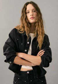 Čierna rifľová bunda so striebornými gombíkmi, s krátkym strihom a vyhrnutými rukávmi, nosená cez svetlý top.