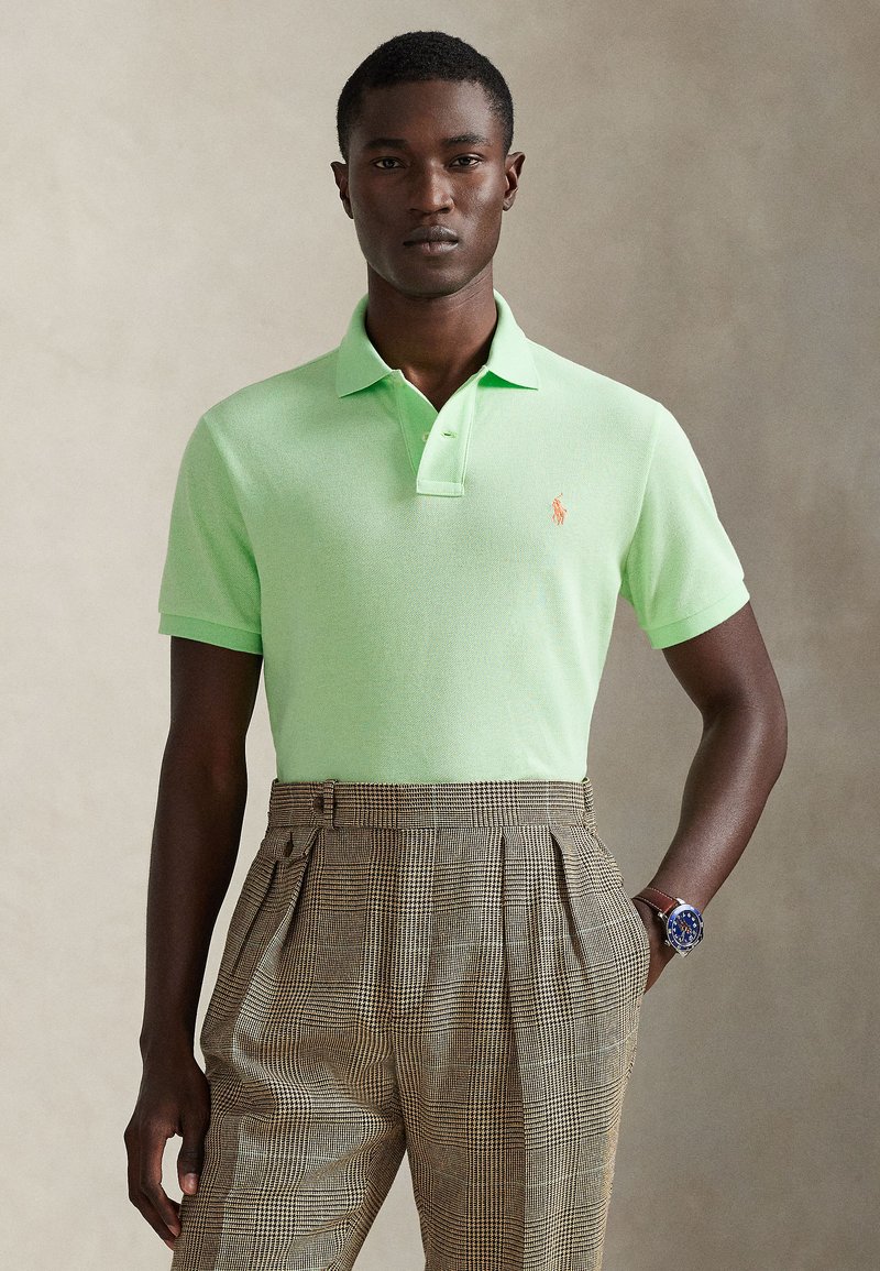 Ανοιχτό πράσινο πόλο μπλουζάκι με  κλασικό γιακά και κοντά μανίκια. Διαθέτει μικρό πορτοκαλί έμβλημα στο αριστερό στήθος. Καφέ καρό παντελόνι.