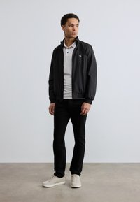 Μαύρο ελαφρύ μπουφάν με ψηλό γιακά και κλείσιμο με φερμουάρ, φορεμένο πάνω από ανοιχτό γκρι πόλο, σε συνδυασμό με μαύρο τζιν και κρεμ παπούτσια. Μινιμαλιστικός σχεδιασμός.