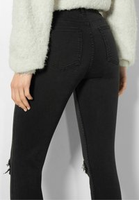 Jeans negros de cintura alta con un diseño ajustado, que presentan detalles desgastados en las rodillas y dos bolsillos traseros, combinados con un suéter blanco de terciopelo.