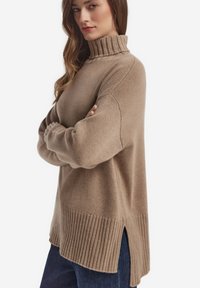 Beige stickad tröja med hög krage, ribbad nederkant och slitsar i sidorna. Designen har en avslappnad passform och mjuk textur.