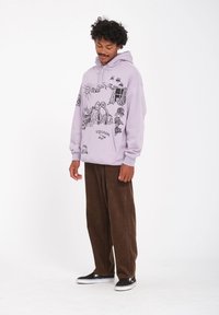 Un sweat à capuche lavande avec des illustrations graphiques noires, comprenant des paysages et des figures, associé à un pantalon en velours côtelé marron et des baskets noires.