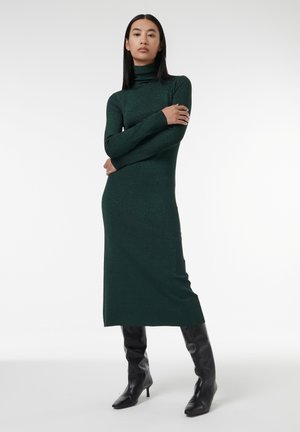 Groene, lange mouw, coltrui jurk met een aansluitende silhoutte en subtiele glans; gedragen met zwarte, kniehoge, haklaarzen.