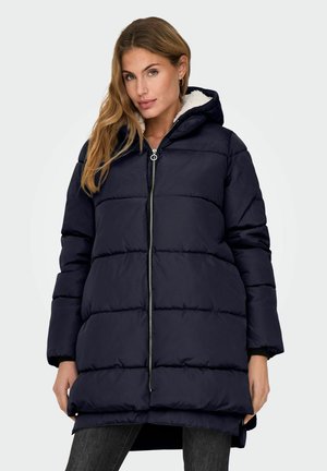 Femme portant un long manteau matelassé bleu marine avec une capuche en polaire blanche et une fermeture éclair à l'avant, se tenant contre un fond uni.