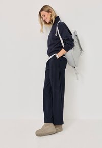 Navy pullover with high collar, gray accents; pinstriped dark pants; beige slip-on shoes; gray backpack; minimalistic design.