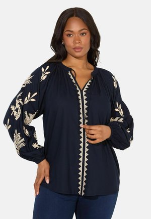 Marinebluse med lange pufærmer. Har beige blomsterbroderi på ærmerne og en dekorativ halsudskæring. Letvægtsstof, afslappet pasform.