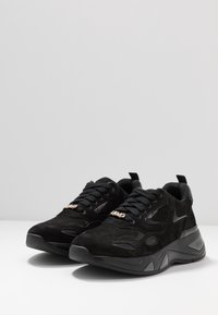 Un paio di sneakers atletiche nere con suole testurizzate, pannelli in rete e suède, lacci neri e dettaglio del marchio metallico sulla linguetta.