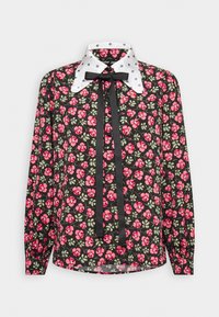 Blouse noire à manches longues avec imprimé floral rose, col blanc à pois noirs et nœud en ruban noir au niveau du cou.