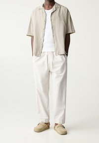 Homme portant une chemise à manches courtes beige à boutons, un débardeur blanc, un pantalon ample blanc à rayures, des chaussures à enfiler beige et un collier en chaîne argenté.