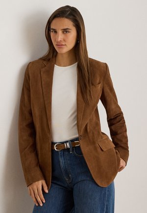 Femme aux longs cheveux bruns porte un blazer en daim marron, une chemise blanche, un jean bleu et une ceinture marron, debout contre un mur clair.