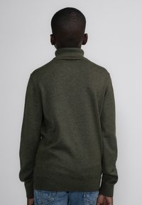 Maglione verde lavorato a maglia con colletto alto, maniche lunghe e orlo a costine. Indossato con jeans in denim azzurro chiaro, visto da dietro.