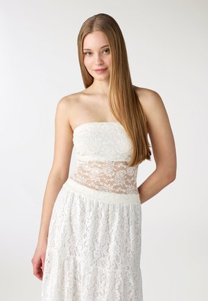 Giovane donna con capelli lunghi e lisci che indossa un abito bianco in pizzo senza spalline, in piedi contro uno sfondo chiaro e semplice.