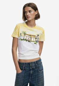 Kortärmad t-shirt med vit kropp och gula gradientstrimmor, med en grafisk design av ett tropiskt hus och palmträd i grönt och brunt.