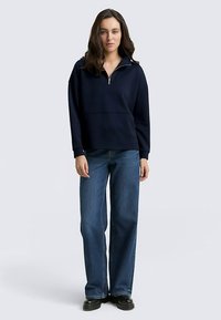 Mørkeblå sweatshirt med kvart lynlås, løs pasform og ribbet tekstur, kombineret med brede blå jeans og chunky sorte sko.