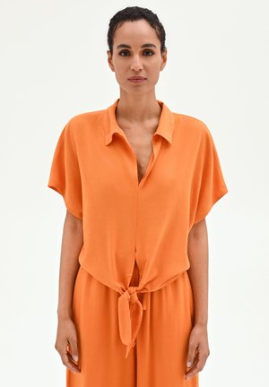 Donna con capelli corti e ricci che indossa una camicia arancione a maniche corte ampia legata in vita e pantaloni larghi abbinati, in piedi davanti a uno sfondo bianco.