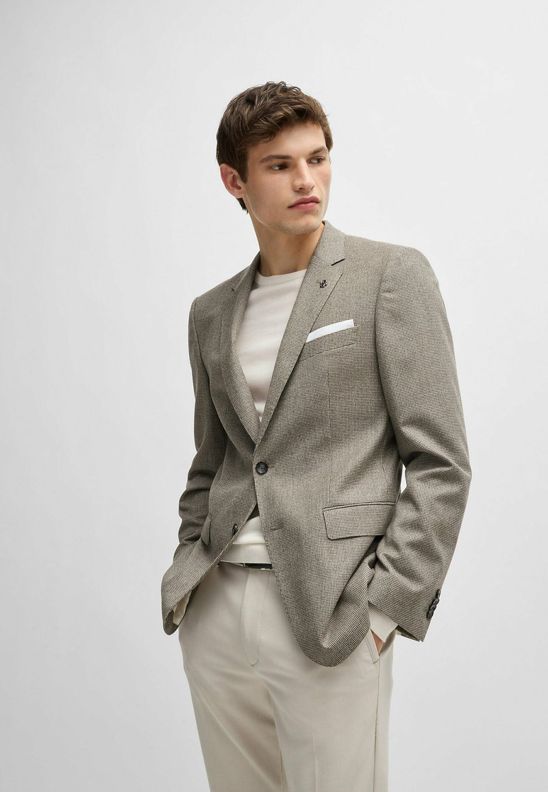 Hellgrauer Blazer mit strukturiertem Muster, Reverskragen, Ein-Knopf-Verschluss und Vordertaschen. Getragen über einem cremefarbenen Hemd und hellen Hosen.