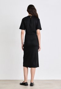 Abito nero con maniche corte, silhouette aderente, lunghezza al ginocchio, dettaglio a chiave sul retro e texture del tessuto liscia. Indossato con ballerine nere.