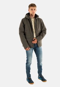 Giacca grigia con zip e cappuccio, due tasche frontali, abbinata a jeans azzurri e scarpe sportive blu con dettagli marroni.