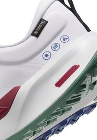 Chaussure de sport blanche avec étiquette Gore-Tex, icônes bleues pour la durabilité, bande rouge sur la semelle intermédiaire et semelle extérieure texturée verte au talon.
