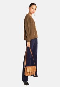 Brauner Strickpullover, marineblaue weit geschnittene Hose und eine goldene metallische Umhängetasche mit Klappe. Die Accessoires umfassen lange goldene Ohrringe und eine Halskette.