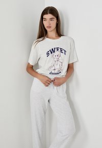 Jonge vrouw die poseert in een lichtgrijze loungewear met een teddybeer en de tekst "SWEET dreams" op het shirt, tegen een plain muur.