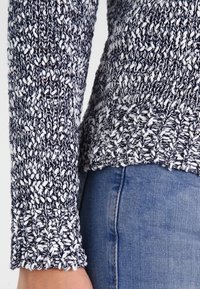 Neulepusero, jossa on laivastonsinisen ja valkoisen langan sekoitus, teksturoitu kuvio, pitkät hihat ja hieman väljä istuvuus vyötäröllä.