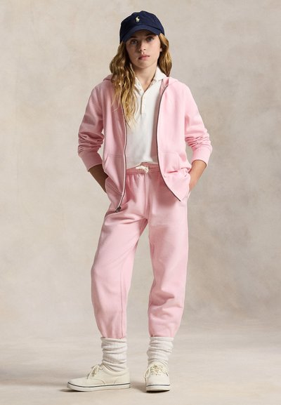 Tânăr stând cu mâinile în buzunare, purtând trening roz, polo alb, adidași albi și șapcă bleumarin cu logo.