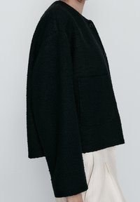 Giacca nera con trama, collo rotondo, due bottoni e tasca frontale. Vestibilità ampia con maniche larghe, indossata sopra pantaloni chiari.