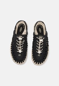 Sorte og beige sandaler med et vævet overdelsdesign, der har hvide snørebånd og en tekstureret sål. Anatomisk fodsengslabel synlig indeni.