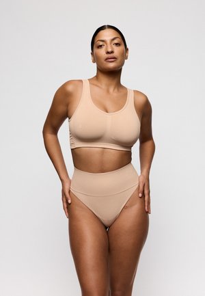 Vrouw met donker haar in een beige naadloze sportbeha en hoge taille ondergoed, staand tegen een effen lichte achtergrond.