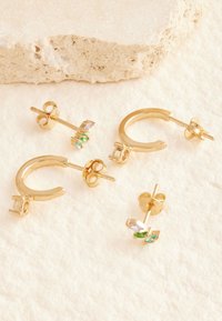 Zwei Paar goldene Ohrringe auf einer beigen, strukturierten Oberfläche, ein Paar mit farbigen Edelsteinen und ein Paar goldene Creolen mit klaren Steinen.